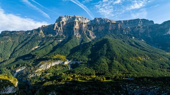 Berglandschaft mit steilen Felswänden und dicht bewaldeten Hügeln unter blauem Himmel.