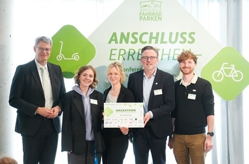 Gruppe von fünf Personen bei der Preisverleihung des Hackathons „Anschluss erreichen“ vor einer Veranstaltungswand. In der Mitte wird die Siegerurkunde präsentiert, im Hintergrund sind das Veranstaltungslogo sowie Symbole für nachhaltige Mobilität zu sehen.<br />
