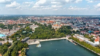 Luftaufnahme einer Stadt mit zentralem Park und Kirche, umgeben von dichter Bebauung und einem See im Vordergrund.