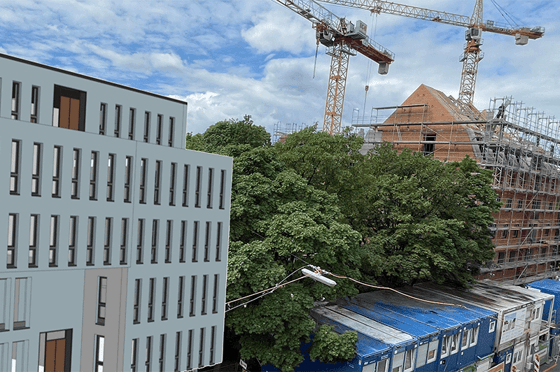 Moderner Gebäudekomplex neben einer Baustelle mit Krananlagen und eingerüstetem Ziegelbau, teilweise von Bäumen verdeckt, unter bewölktem Himmel.
