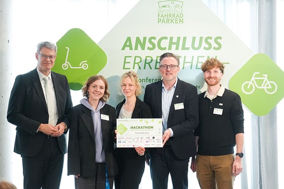 Vier Personen stehen bei der Preisverleihung des DB InfraGO Hackathons „Anschluss erreichen“ auf einer Bühne und präsentieren eine Urkunde vor einer Veranstaltungswand mit grünem Veranstaltungslogo.