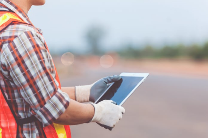 Person in Warnweste und Schutzhandschuhen nutzt ein Tablet auf einer Baustelle oder Vermessungsfläche im Freien. Das mobile Gerät wird für die digitale Datenerfassung, Inspektion oder das Field Mapping vor Ort verwendet. Der Hintergrund ist unscharf und zeigt eine offene Arbeitsumgebung.