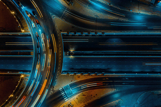 Nächtliche Luftaufnahme eines beleuchteten Autobahnkreuzes mit mehreren Fahrspuren und Lichtspuren fahrender Fahrzeuge. Die sich kreuzenden Verkehrswege symbolisieren Verbindungen, Abhängigkeiten und Netzwerke zwischen verschiedenen Elementen.