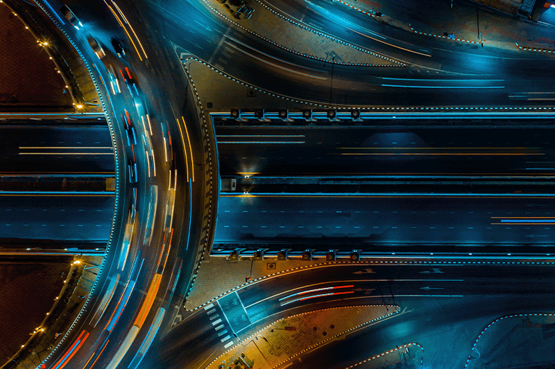 Nächtliche Luftaufnahme eines beleuchteten Autobahnkreuzes mit mehreren Fahrspuren und Lichtspuren fahrender Fahrzeuge. Die sich kreuzenden Verkehrswege symbolisieren Verbindungen, Abhängigkeiten und Netzwerke zwischen verschiedenen Elementen.