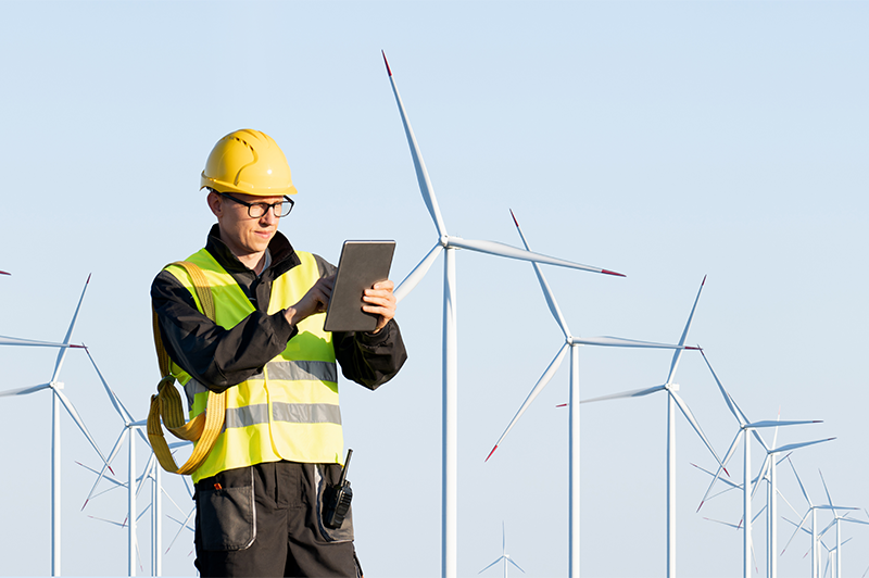 Person mit Schutzhelm und Warnweste nutzt ein Tablet vor mehreren Windenergieanlagen in einer offenen Landschaft.