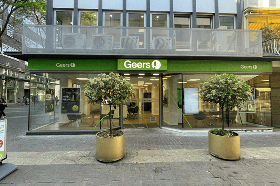 Eingangsbereich eines modernen Geschäfts mit der grünen Beschilderung „Geers“. Große Glasfronten geben den Blick ins helle Innere frei, in dem Sitzbereiche und Displays zu sehen sind. Vor dem Eingang stehen zwei runde, goldfarbene Pflanzkübel mit kleinen Bäumen. Das Geschäft befindet sich in einem Gebäude mit Balkon darüber, auf dem mehrere Klimaanlagen installiert sind. Links und rechts des Eingangs führen Gehwege entlang einer belebten Straße mit weiteren Gebäuden und Passantinnen und Passanten.