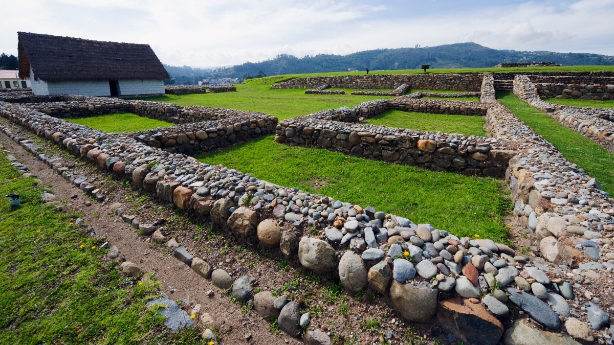 Archäologische Steinanlage mit niedrigen Mauern aus runden Steinen, umgeben von grüner Landschaft und einem Haus mit Strohdach.