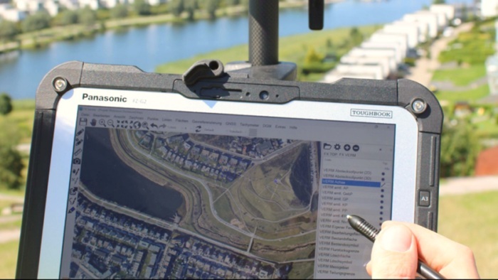Tablet mit GIS‑Kartenanwendung im Außeneinsatz, auf dem Luftbilder und Geodaten dargestellt und per Stift bearbeitet werden.