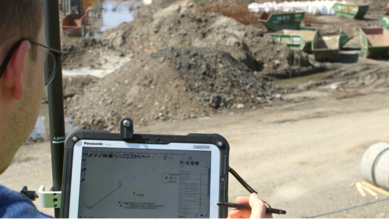 Person im Außeneinsatz arbeitet mit einem Tablet und Stift an einer GIS‑Kartenanwendung auf einer Baustelle mit Erd- und Materialhaufen im Hintergrund.