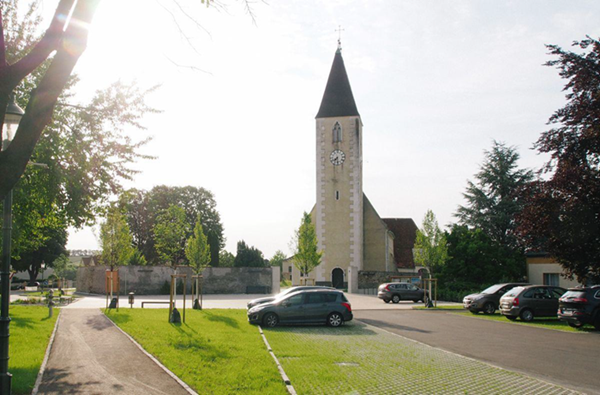Aufnahme eines ruhigen Dorf- oder Kleinstadtbereichs mit einer Kirche im Zentrum. Die Kirche besteht aus einem hellen Turm mit spitzem Dach und Uhr sowie einem angrenzenden Kirchengebäude mit Ziegeldach. Im Vordergrund befindet sich ein Parkplatz mit mehreren abgestellten Autos auf teilweise begrüntem Pflaster. Links verläuft ein Fuß- und Radweg entlang einer Grünfläche mit jungen Bäumen. Im Hintergrund sind weitere Gebäude, Bäume und eine Mauer sichtbar. Die Szene ist von hellem Tageslicht geprägt, mit leichtem Gegenlicht durch die Sonne oben links.