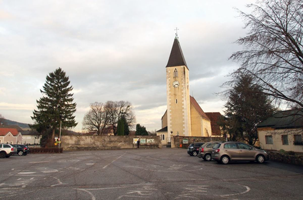 Aufnahme eines Dorf- oder Kleinstadtplatzes mit einer Kirche im Hintergrund. Die Kirche hat einen hohen, hellen Turm mit spitzem Dach und Uhr sowie ein angrenzendes Gebäude mit rotem Ziegeldach. Im Vordergrund erstreckt sich ein großer, grauer Asphaltparkplatz mit eingezeichneten Markierungen und mehreren parkenden Autos am Rand. Links steht ein großer Nadelbaum, daneben eine niedrige Mauer, die den Platz von dahinterliegenden Gebäuden trennt. Rechts sind weitere Bäume ohne Blätter sowie ein kleines Gebäude zu sehen. Der Himmel ist bewölkt, und die Szene wirkt insgesamt kühler und grauer als eine typische Sommerszene.