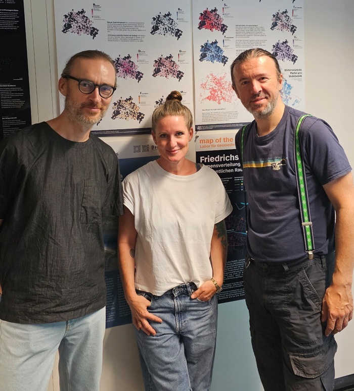 Drei Personen stehen vor wissenschaftlichen Postern mit Kartenvisualisierungen und Geodaten-Diagrammen in einem Ausstellungs- oder Forschungsumfeld.