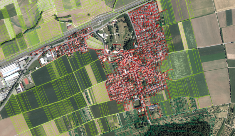 Luftbildbasierte Kartenansicht eines ländlichen Ortes mit rot hervorgehobenen Gebäude- und Straßengeometrien im Ortskern sowie grün umrandeten landwirtschaftlichen Parzellen im Umland; Felder, Wege und Siedlungsstrukturen sind deutlich voneinander abgegrenzt.