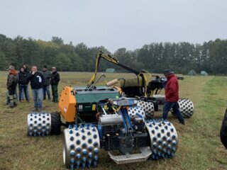 Auf einer offenen Wiesen‑ oder Moorfläche steht ein großes landwirtschaftliches Spezialfahrzeug mit mehreren breiten Metallwalzen, die mit blauen Noppen besetzt sind. Das Fahrzeug ist in Grün, Orange und Blau gehalten und verfügt über einen Aufbau mit technischen Komponenten. Im Hintergrund stehen mehrere Personen in wetterfester Kleidung und beobachten die Vorführung. Die Szene spielt bei bewölktem Himmel, umgeben von Bäumen am Rand der Fläche.